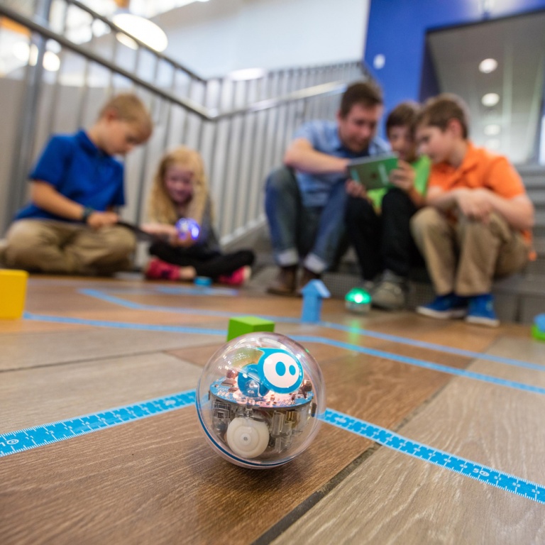 Liten bollformad robot på golv, i bakgrunden barn och en vuxen sittandes på golvet